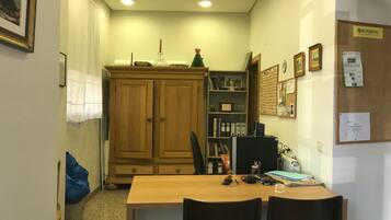 A small office with a wooden desk, two chairs, a bookshelf, and a bulletin board with papers.