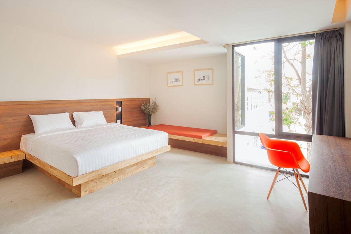 Moderni hotellihuone, jossa on puinen sänky, penkki, suuri ikkuna ja punainen tuoli.