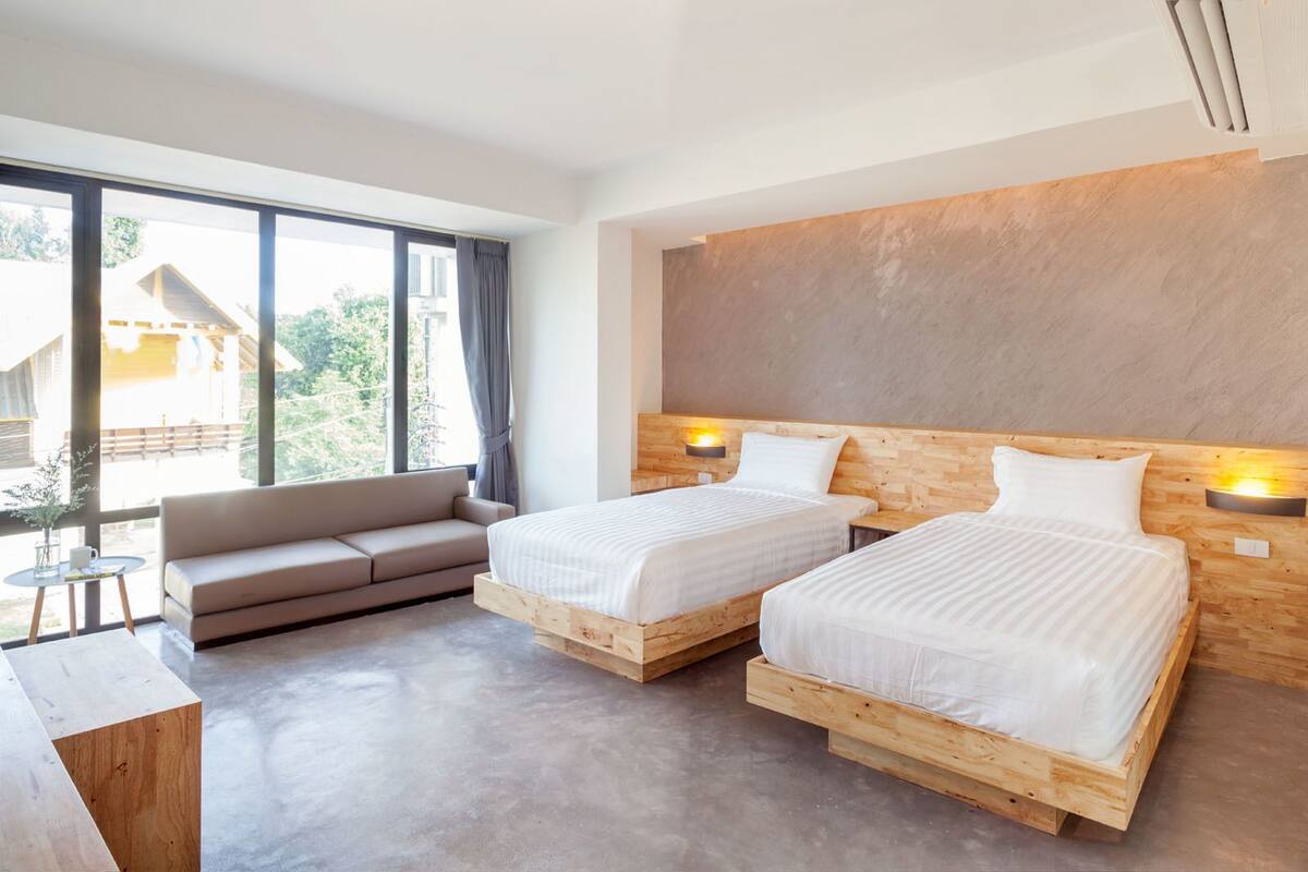 Moderni hotellihuone, jossa on kaksi puista sänkyä, sohva ja suuri ikkuna, josta avautuu näkymä ulos.