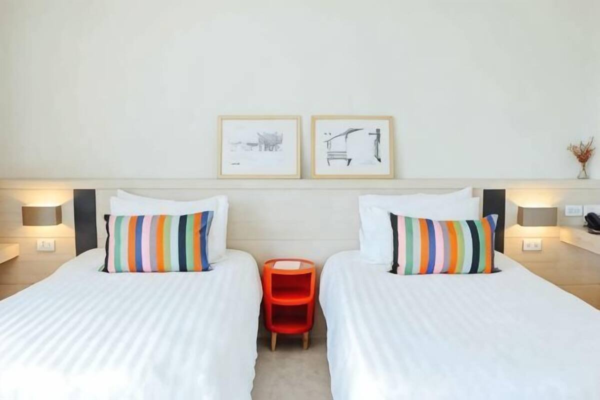 Hotellihuone, jossa on kaksi sänkyä, pieni punainen jakkara ja seinällä kehystetty taideteos.