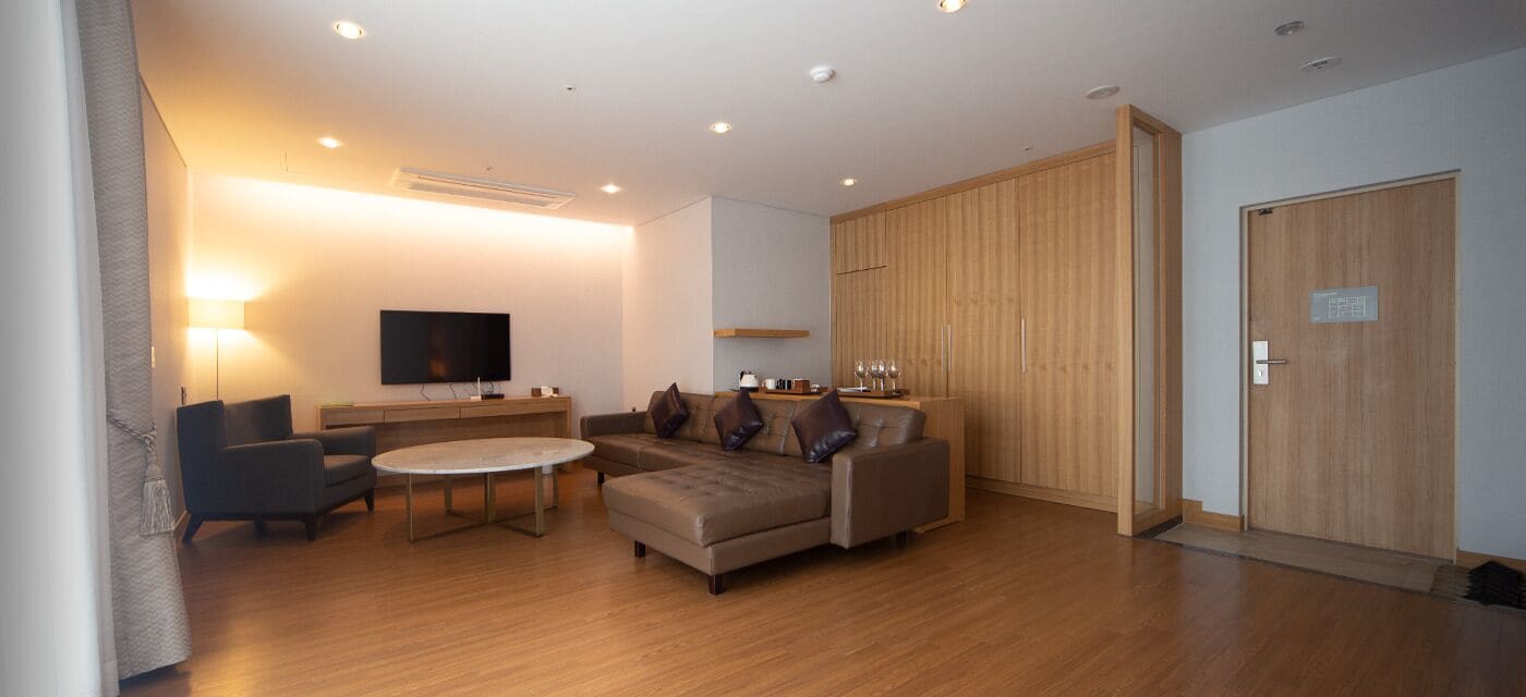 Una sala moderna con un sofá, sillón y una mesita de centro. Hay un televisor montado en la pared, un armario de madera y una puerta con un letrero.