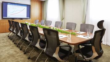 Un salón de conferencias con una mesa larga, varias sillas, una pantalla grande que muestra un gráfico y un ventanal con cortinas.