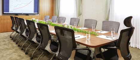 Un salón de conferencias con una mesa larga, varias sillas, una pantalla grande que muestra un gráfico y un ventanal con cortinas.
