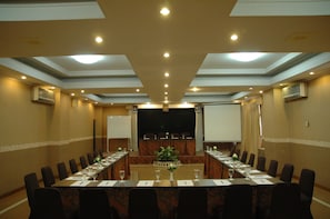 Un salón de conferencias con una mesa larga, sillas, una pantalla de proyector y un escenario.