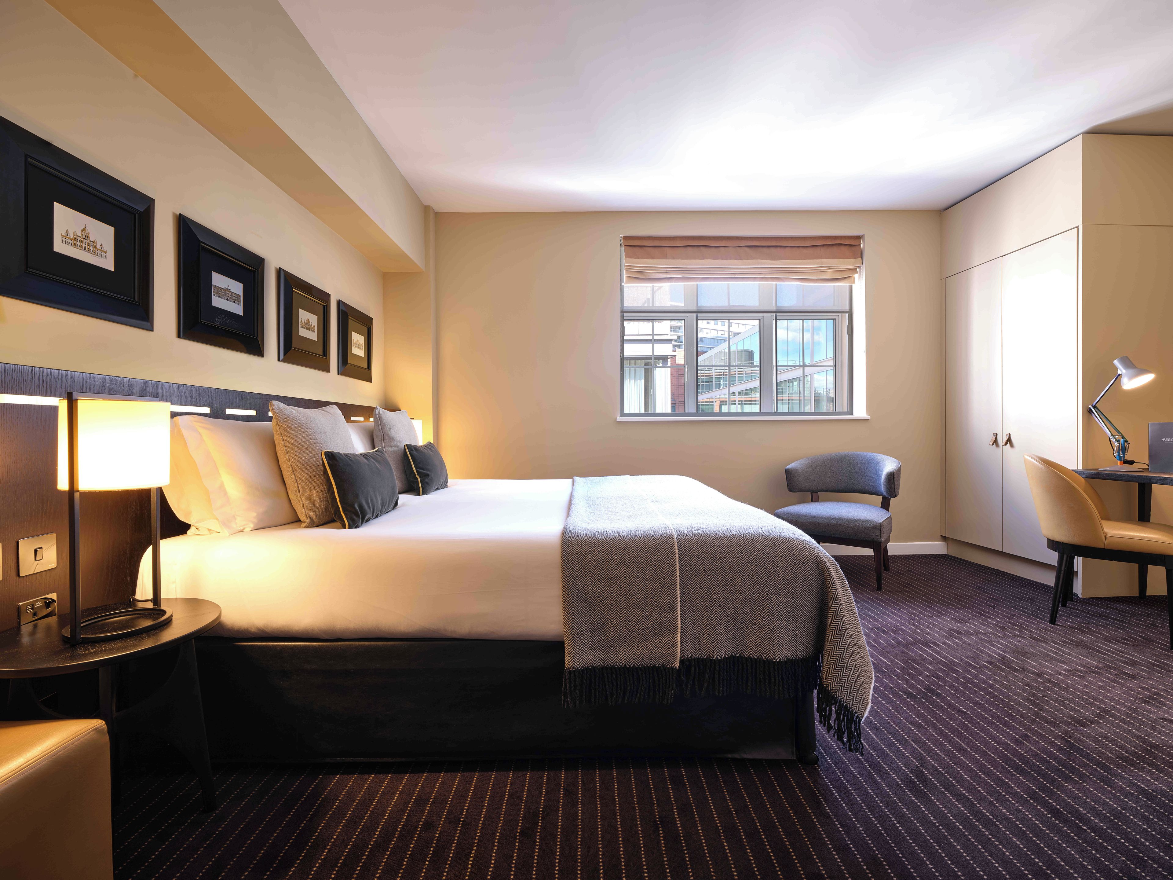 A hotel room with a bed, a desk, a chair, a lamp, and a window with a city view.