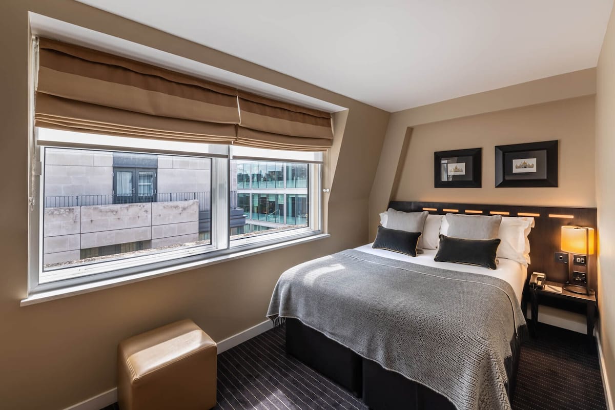A hotel room with a large window, a bed with a grey blanket, a bedside table with a lamp, and two framed pictures on the wall.