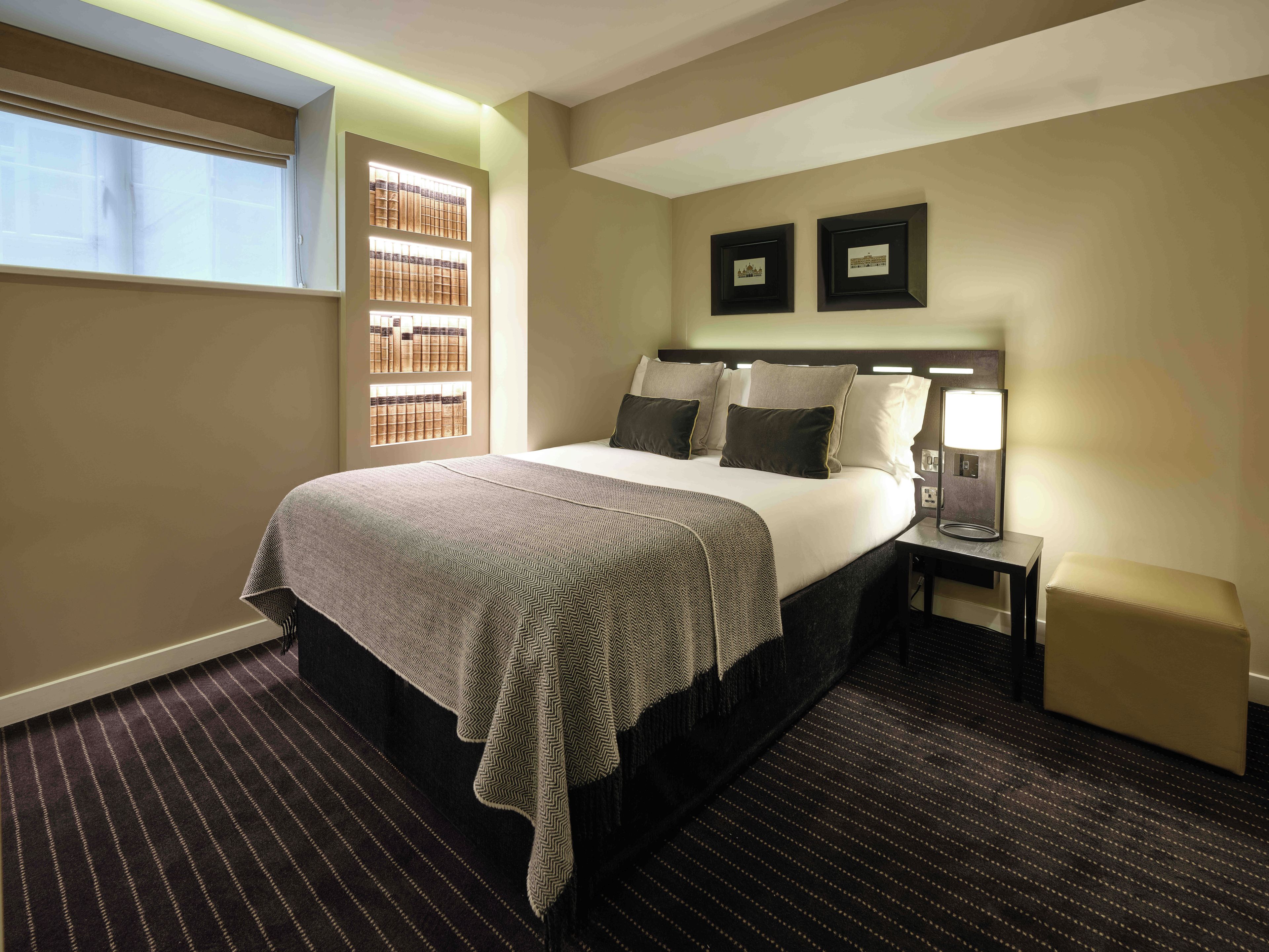 Een hotelkamer met een groot bed, een nachtkastje met een lamp, een klein krukje, een raam met luxaflex en twee ingelijste foto's aan de muur.
