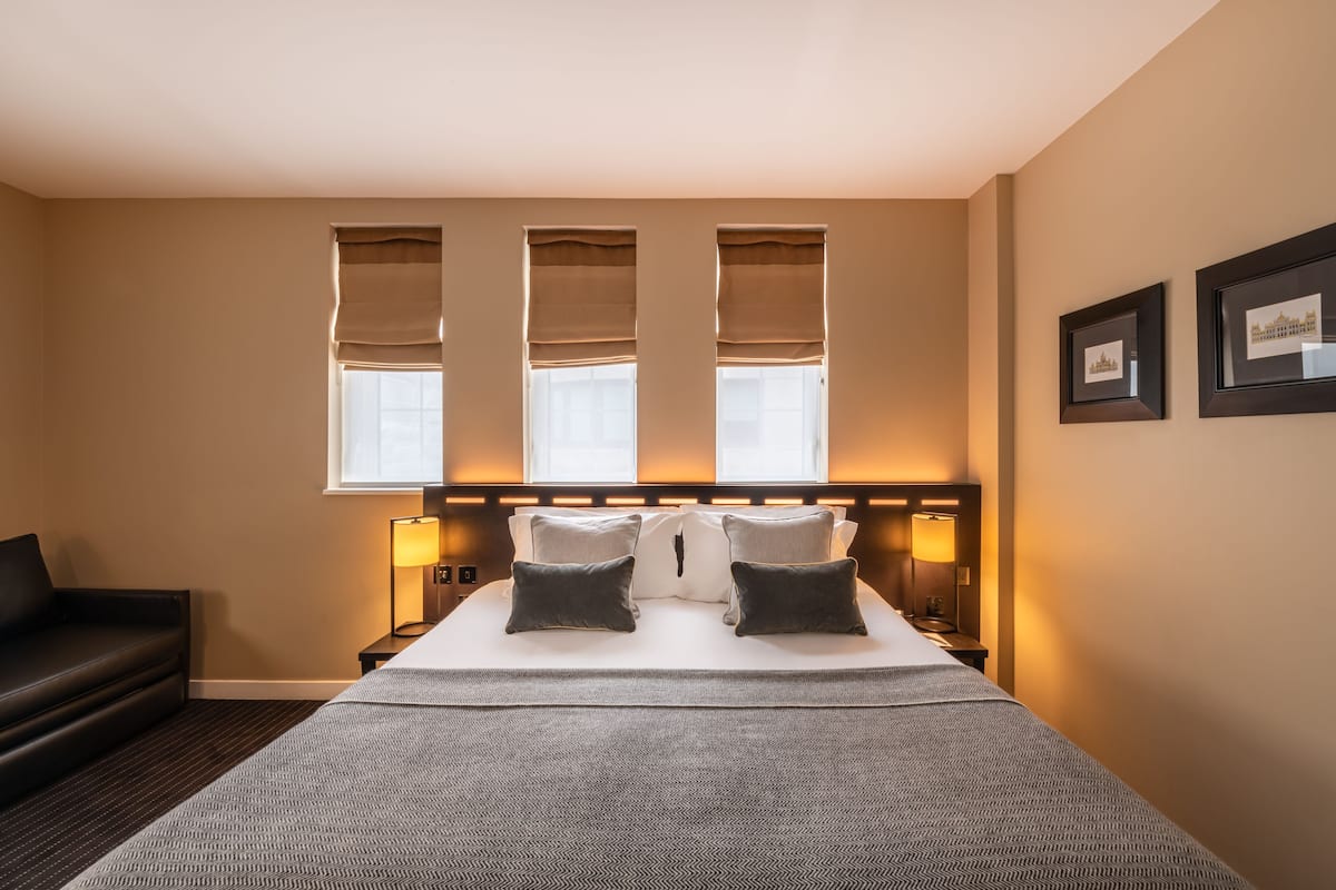A hotel room with a large bed, two bedside lamps, a headboard with built-in lighting, a black leather sofa, and two windows with beige blinds.