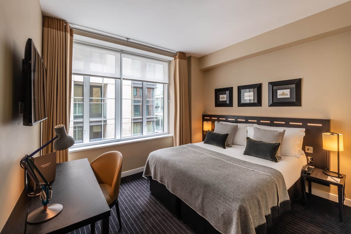 A hotel room with a large bed, a desk with a lamp, a chair, a nightstand with a lamp, and a window with curtains.