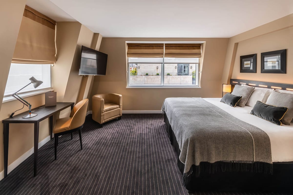 A hotel room with a bed, a desk with a lamp, a chair, a TV mounted on the wall, and a window with blinds.