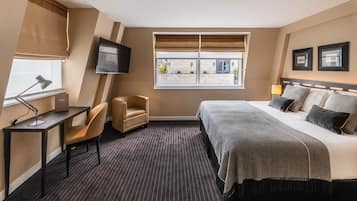 A hotel room with a bed, a desk with a lamp, a chair, a TV mounted on the wall, and a window with blinds.