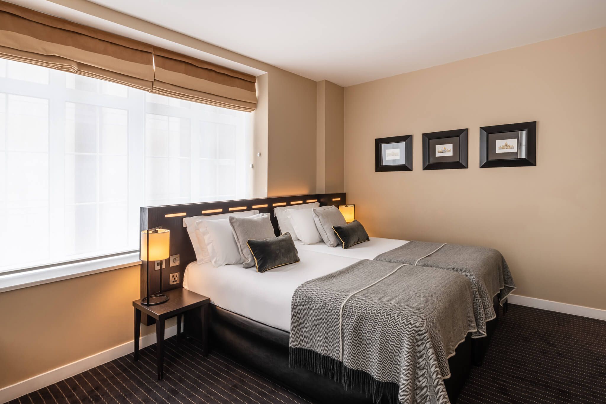 A hotel room with a large bed, a bedside table with a lamp, a window with blinds, and three framed pictures on the wall.