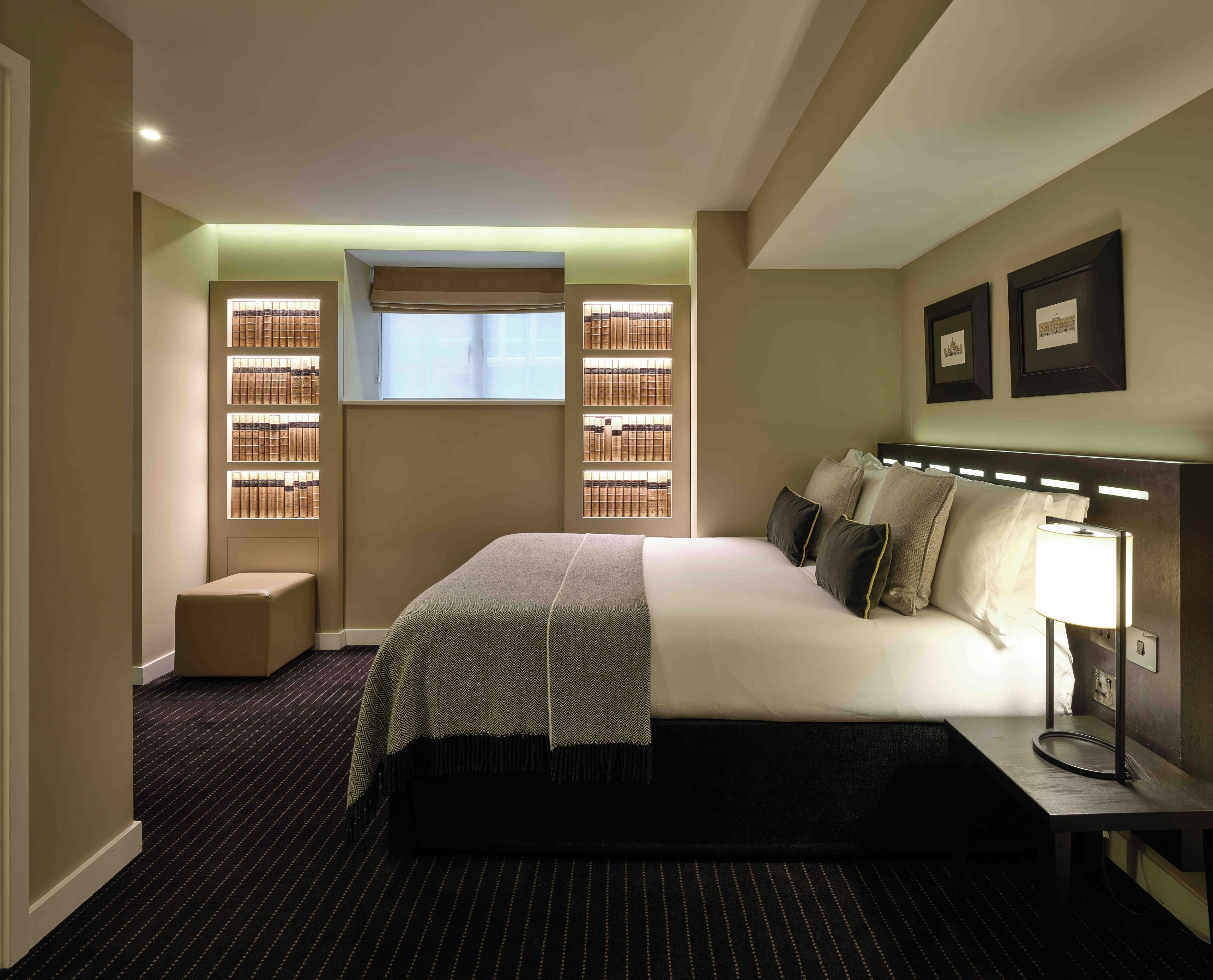A hotel room with a bed, bedside table, lamp, and a window with blinds.