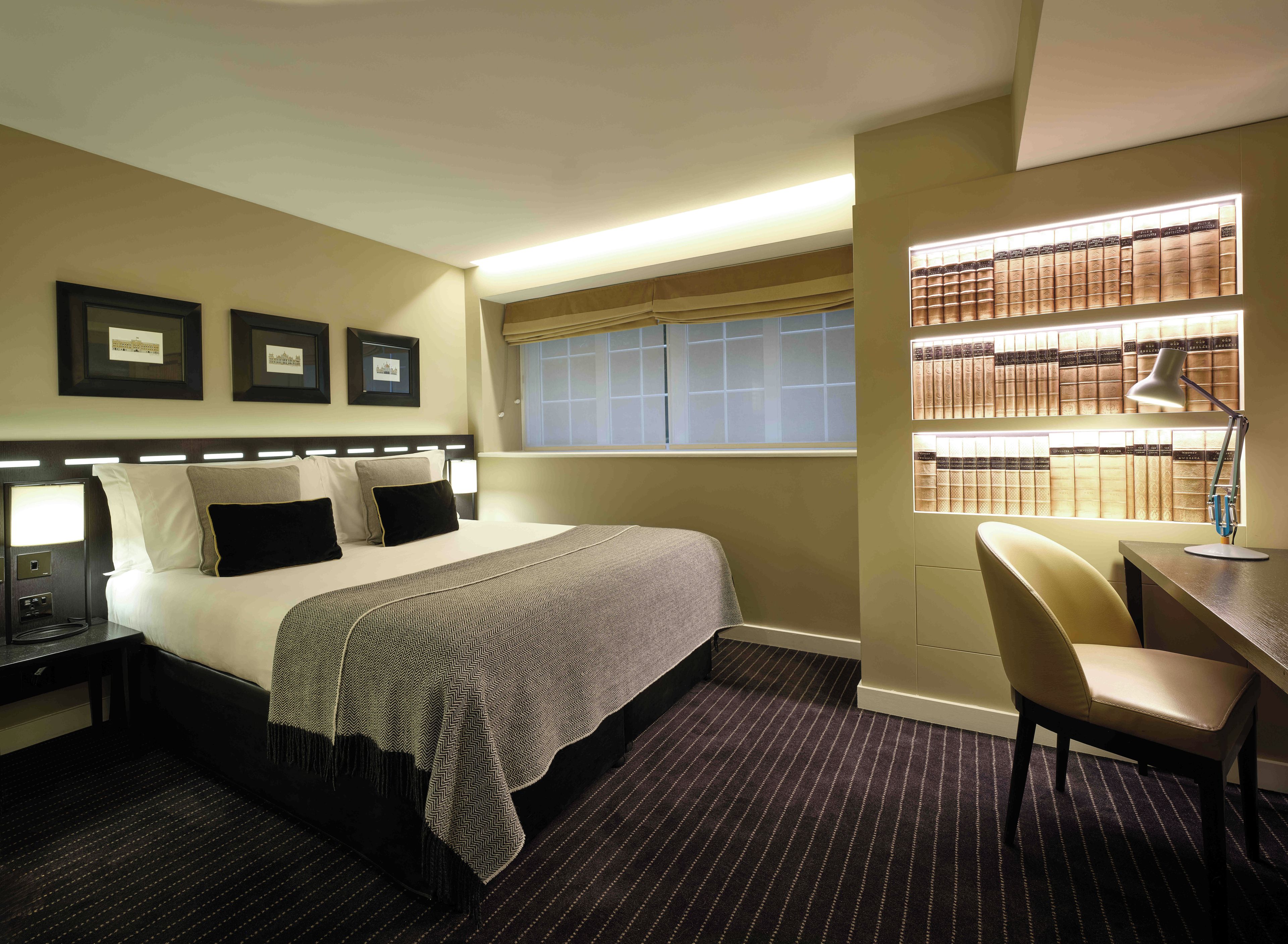 A hotel room with a bed, bedside table, lamp, chair, window, and bookshelf.