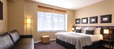 A hotel room with a bed, a sofa, a window with curtains, framed pictures on the wall, and a lamp.