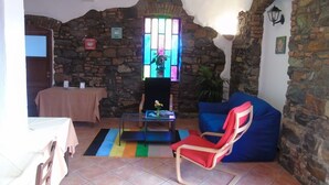 Una habitación rústica con paredes de piedra, una puerta de madera, una mesa con mantel rosa, un sofá azul, una silla roja y una alfombra de rayas coloridas.