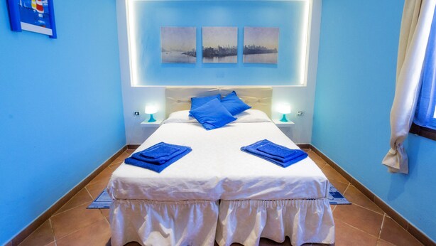 Cama doble con ropa de cama blanca y almohadas azules, mesita de noche con lámpara y tres cuadros colgados en la pared.