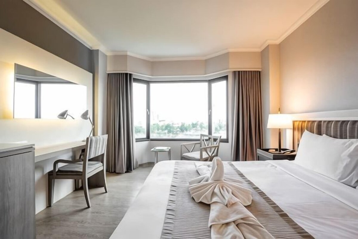 A hotel room with a large bed, a desk, a chair, a nightstand, and a view of the outdoors through a window.
