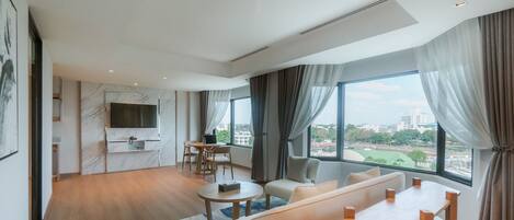 A modern hotel room with a large window, a flat-screen TV mounted on a marble wall, a dining area with a table and chairs, and a sofa.