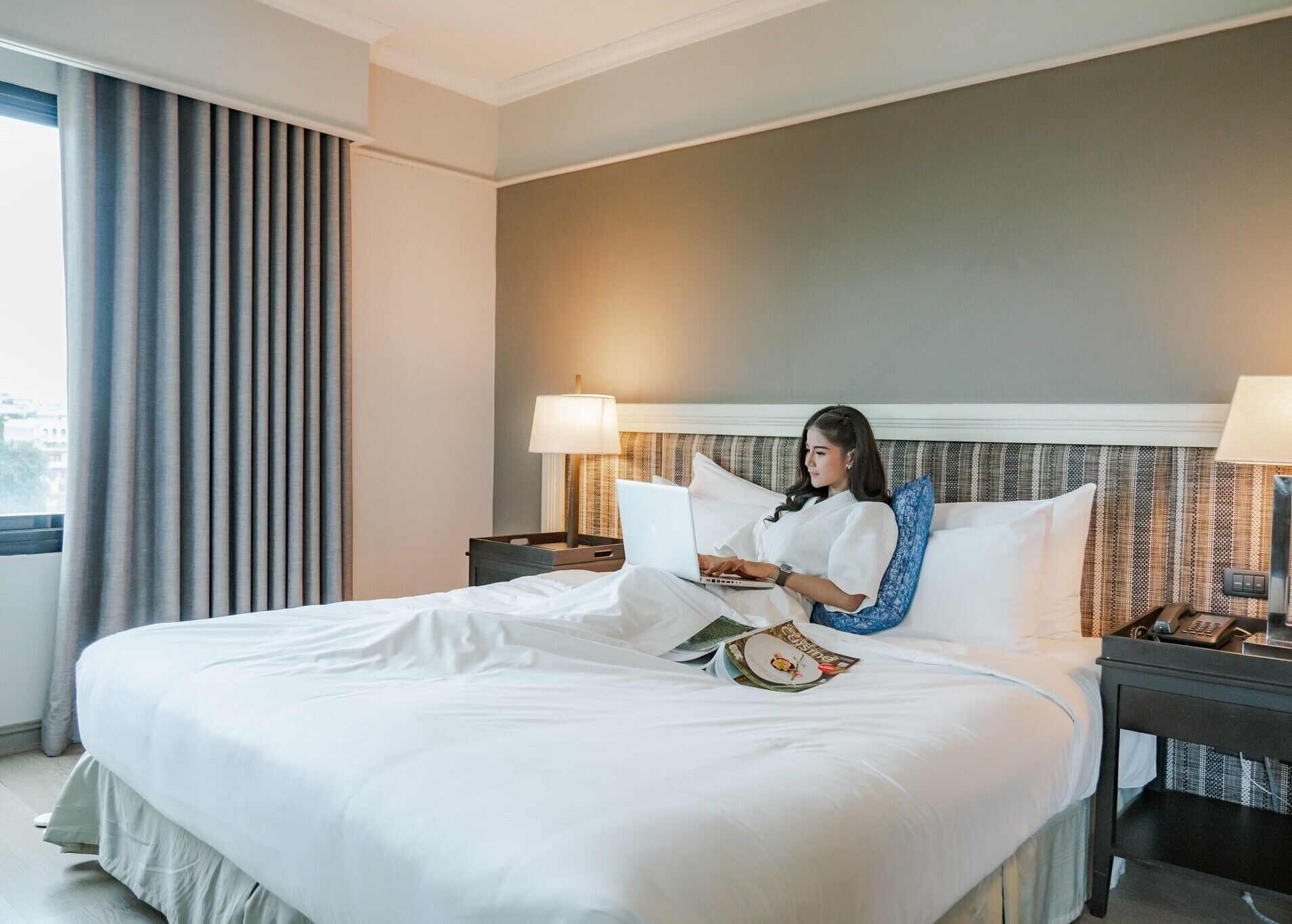A hotel room with a large bed, a laptop, a magazine, and a nightstand with a phone.