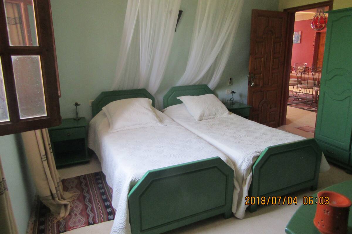 Un lit double avec une tête de lit verte et du linge de lit blanc, dans une pièce dotée d’une porte en bois et d’une petite table avec une lampe.