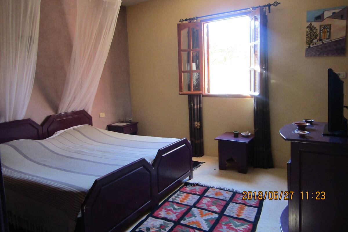 Une chambre à coucher comprenant un lit, une table de chevet, une commode, une fenêtre avec des rideaux et un tableau accroché au mur.