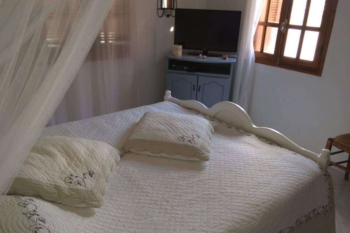 Un lit bien fait, recouvert d’une couette blanche et agrémenté de coussins décoratifs, un téléviseur posé sur un meuble, et une fenêtre avec des rideaux.