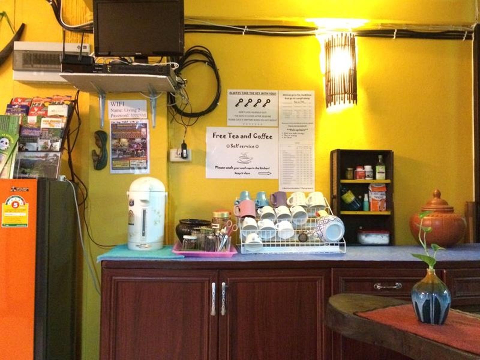 A small cafe with a yellow wall, a coffee machine, a self-service tea and coffee station, and a shelf with various items.