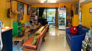 A room with a long wooden bench, a blue sofa, a green bookshelf, and a refrigerator with a 'Cool' sign. - The Living Place 2 - Hostel (Chiang Mai)