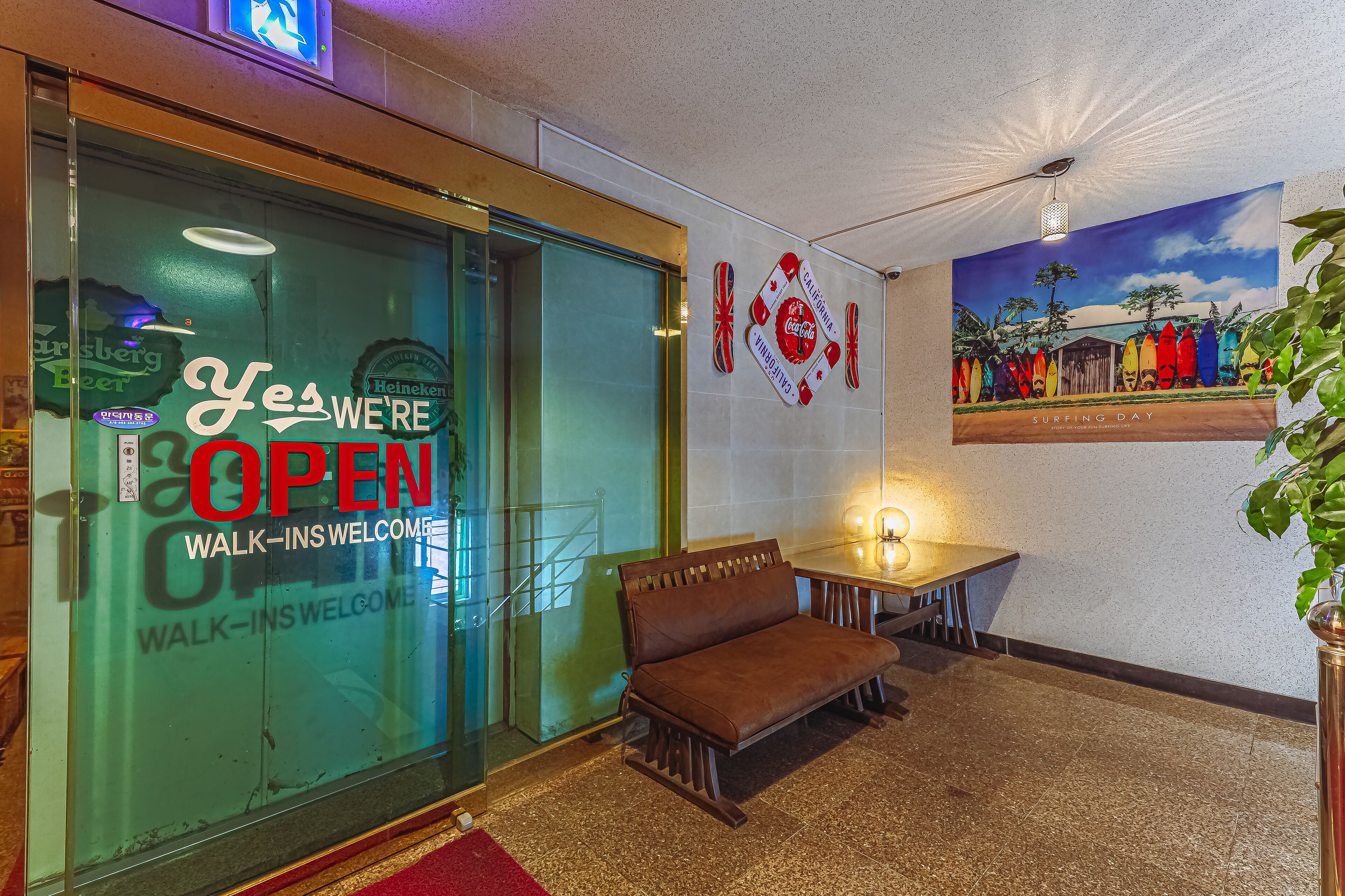 A hotel lobby with a glass door displaying a 'Yes We're Open' sign, a bench, a small table with a lamp, and a surfboard mural on the wall.