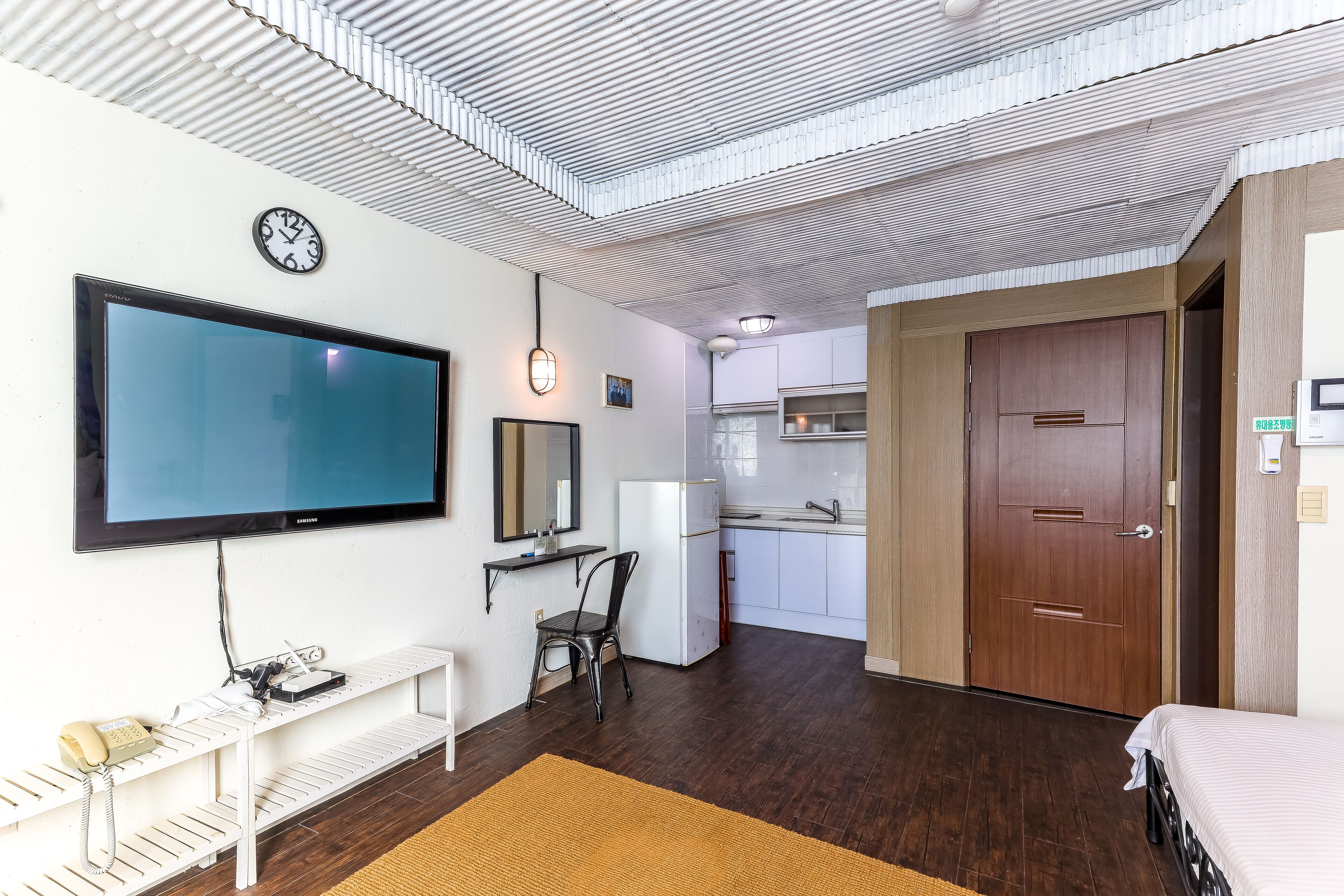 Una habitación de hotel moderna con una televisión de pantalla plana grande, un escritorio con teléfono, una silla y una puerta de madera que da a una zona de cocina.