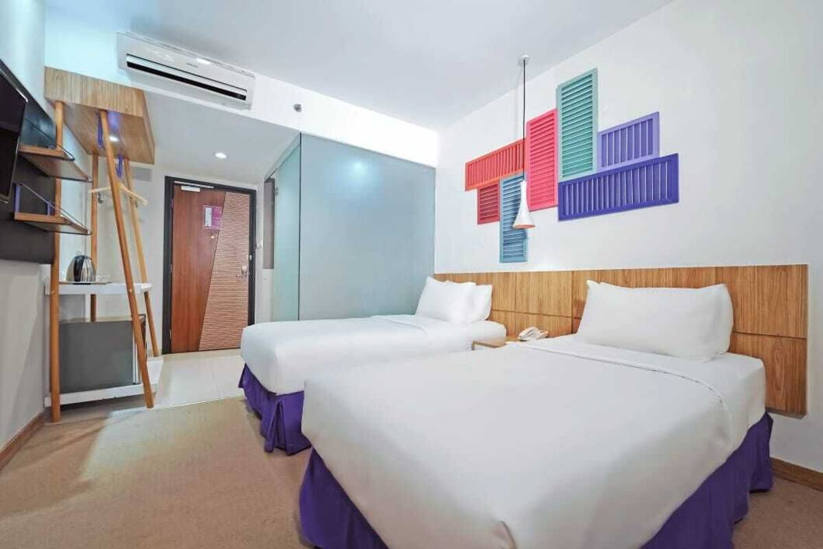 Ett hotellrum med två sängar, en sänggavel i trä, en väggmonterad luftkonditioneringsanläggning och ett färgstarkt, abstrakt konstverk på väggen.