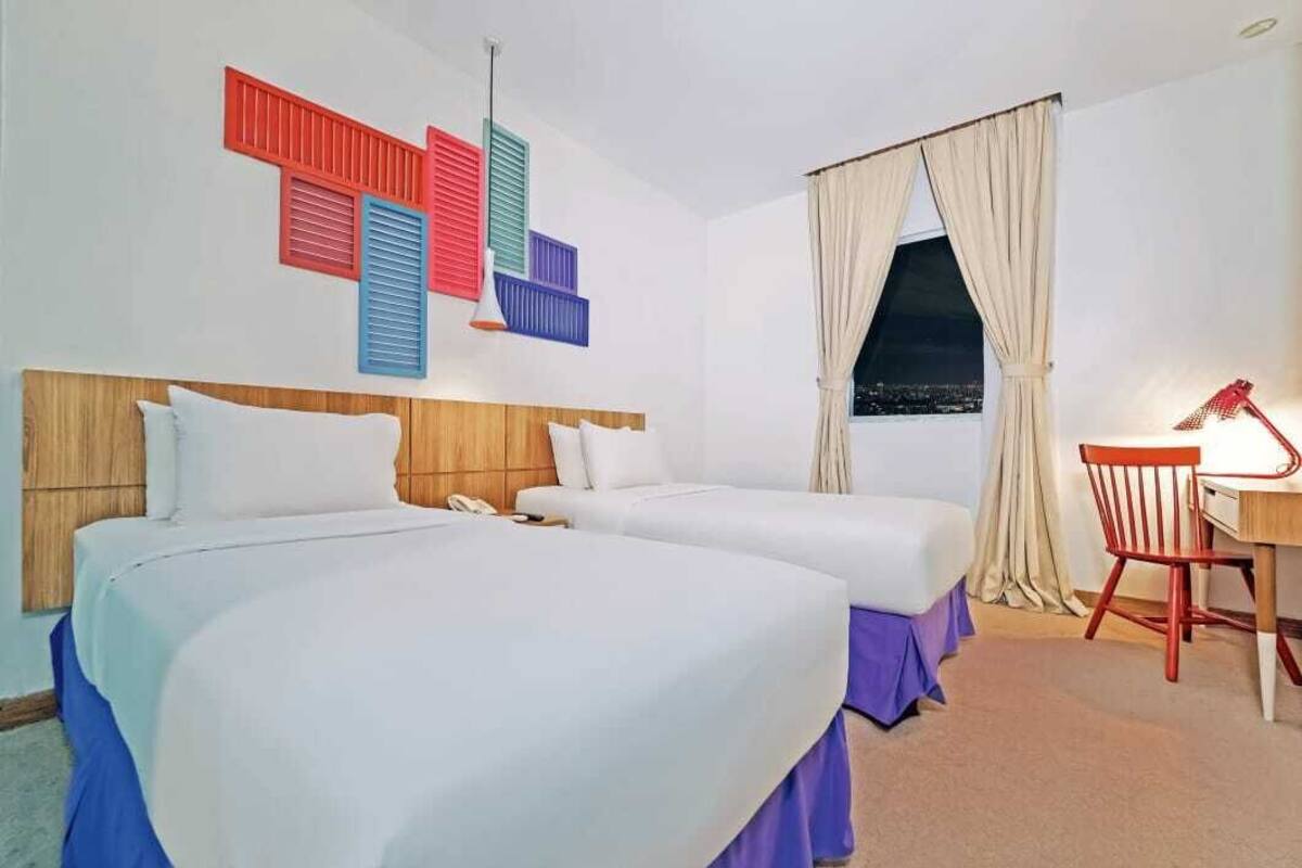 Ett hotellrum med två sängar, ett skrivbord, en stol och ett fönster med gardiner.