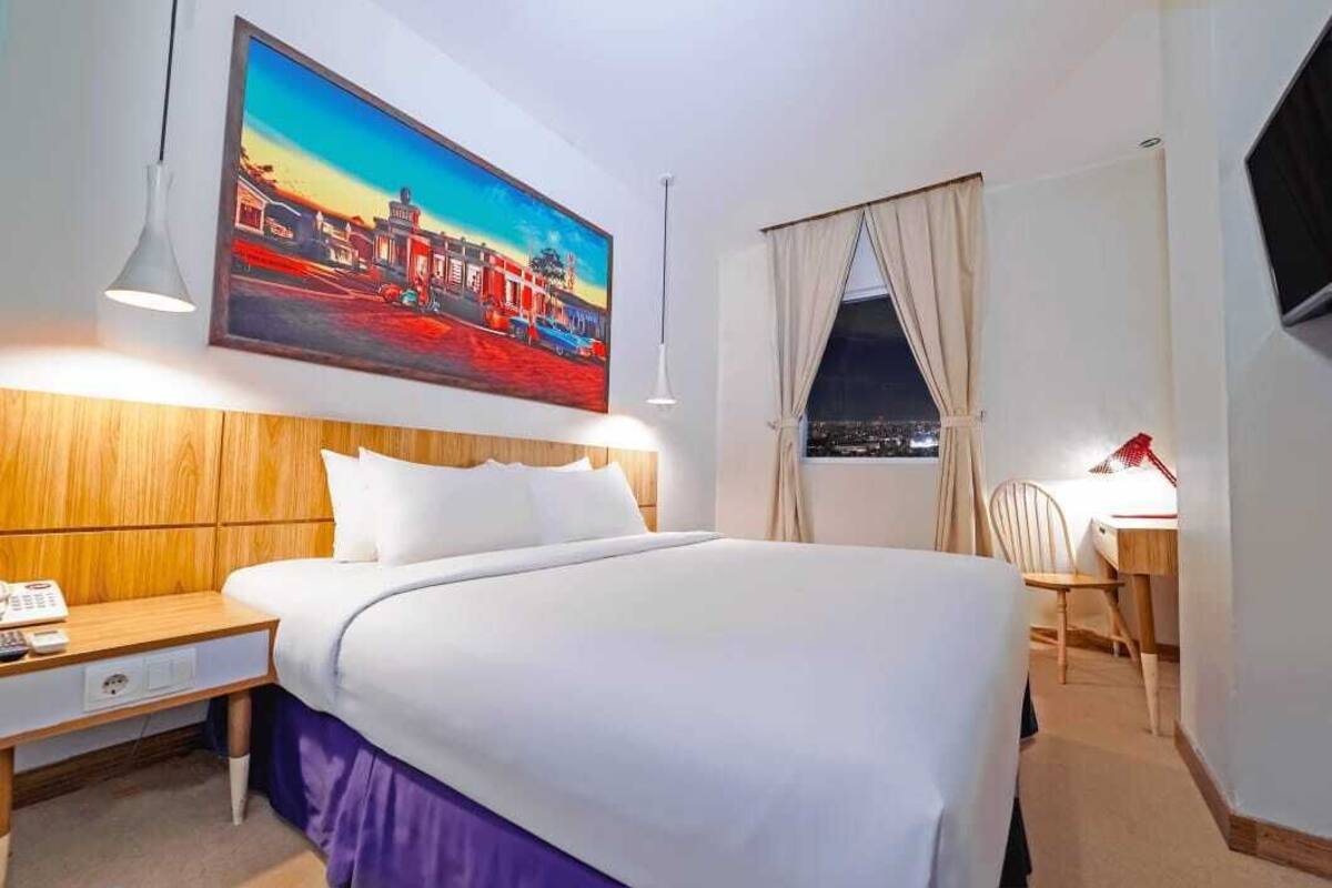 Ett hotellrum med en stor säng, ett skrivbord, en stol, en tavla på väggen och ett fönster med gardiner.