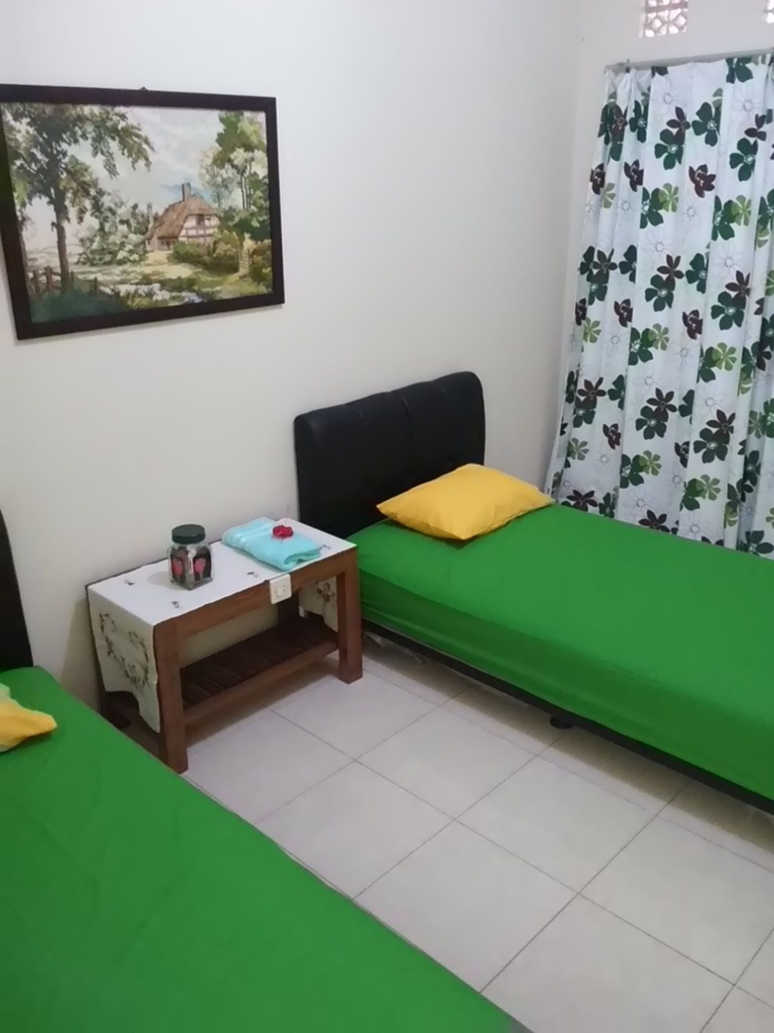 Un dormitorio con una cama individual, una mesita de noche, una pequeña mesa, un cuadro en la pared y una ventana con cortina estampada.