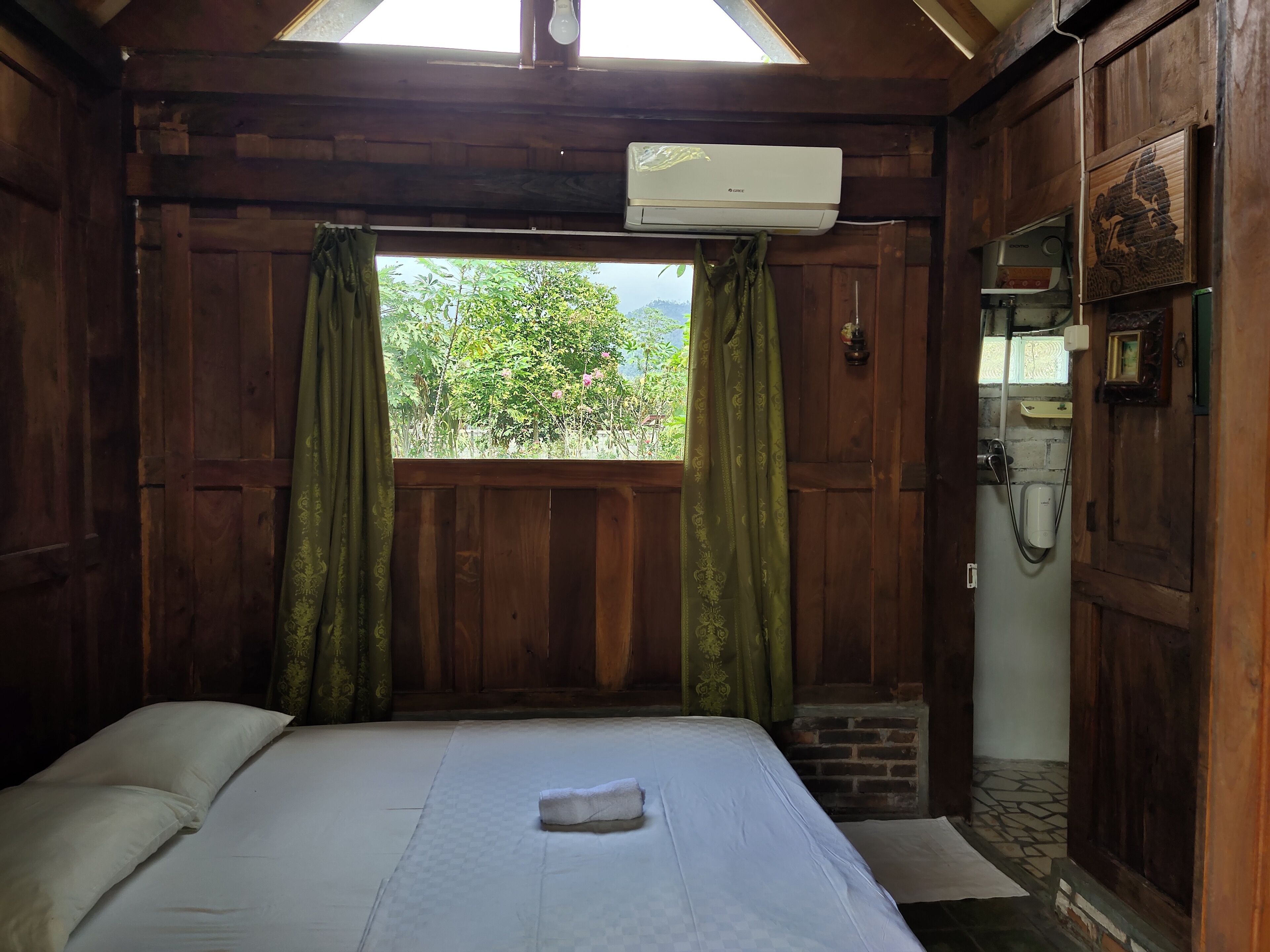 Una habitación con cama, ventana con cortinas, unidad de aire acondicionado y una pequeña estructura de ladrillo.