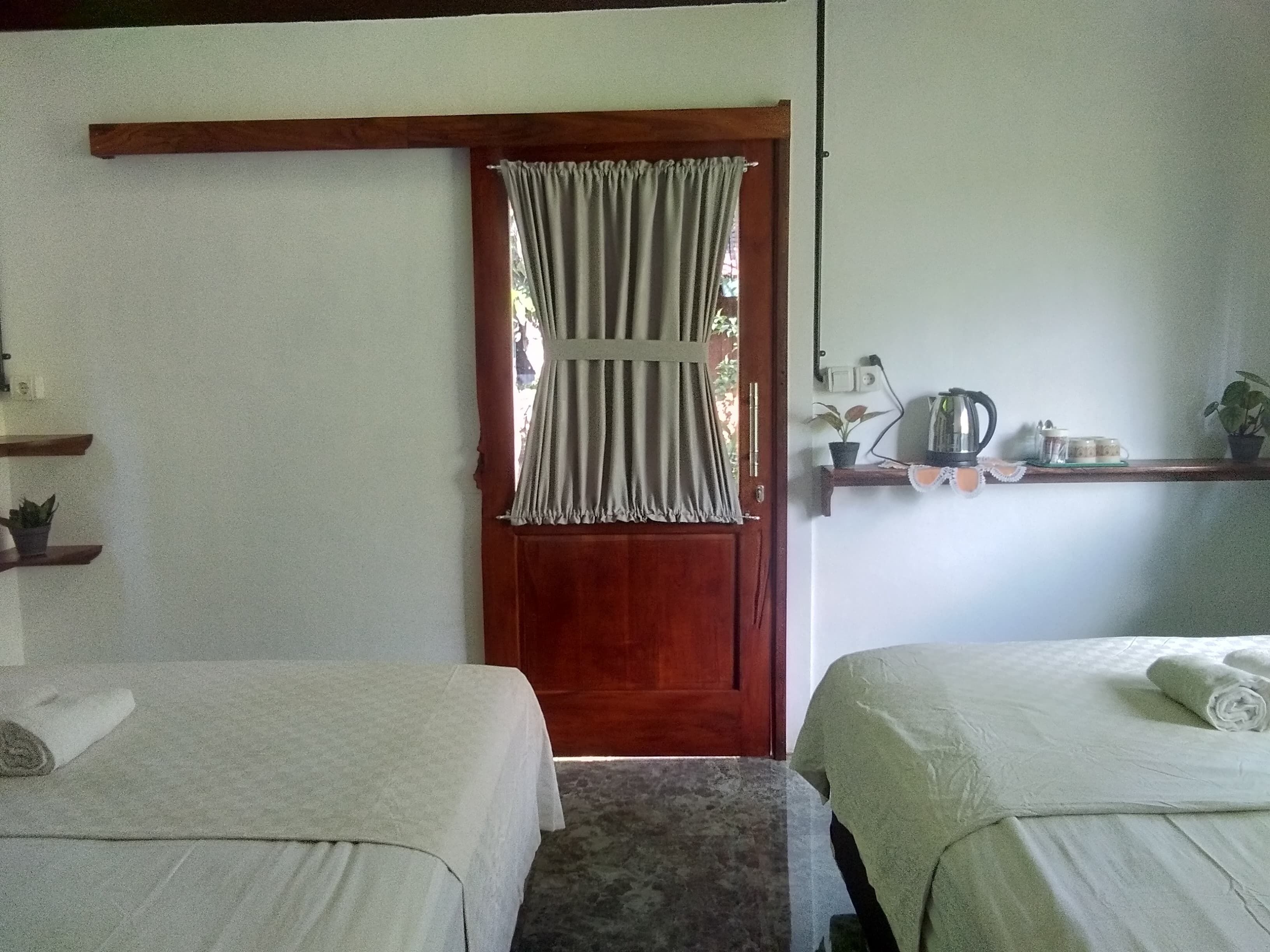A room with two beds, a wooden door, a shelf with a kettle, and a small plant.