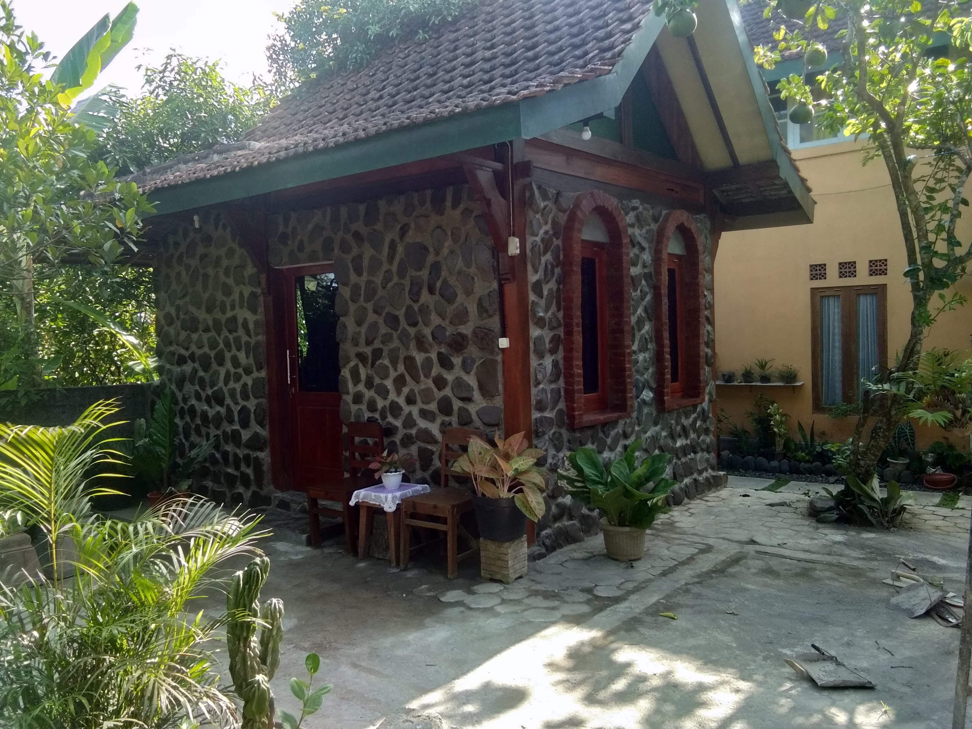 Una casa de piedra con techo de tejas, puerta de madera y ventanas. Hay una mesa con sillas afuera, rodeada de macetas con plantas.