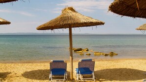 Une plage avec deux chaises longues bleues placées sous des parasols en chaume, une vue dégagée sur la mer et des affleurements rocheux dans l’eau.