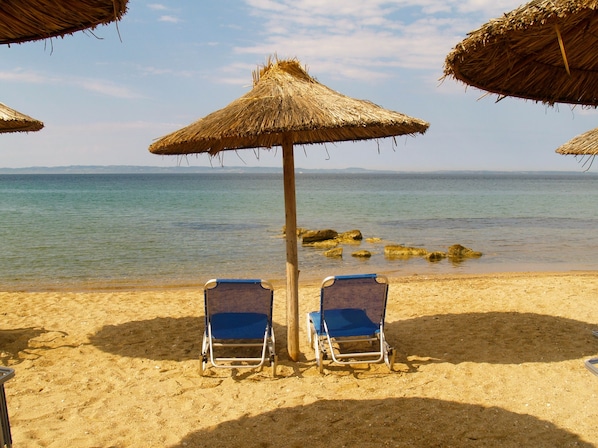 A beach with two blue lounge chairs under thatched umbrellas, a clear view of the sea, and rocky outcrops in the water.