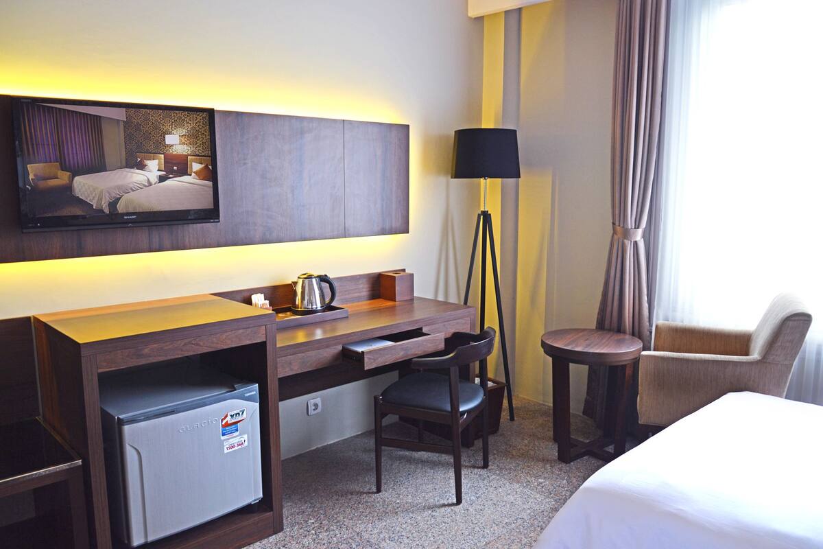 A hotel room with a bed, a desk with a kettle, a chair, a small table, a television, and a minibar.