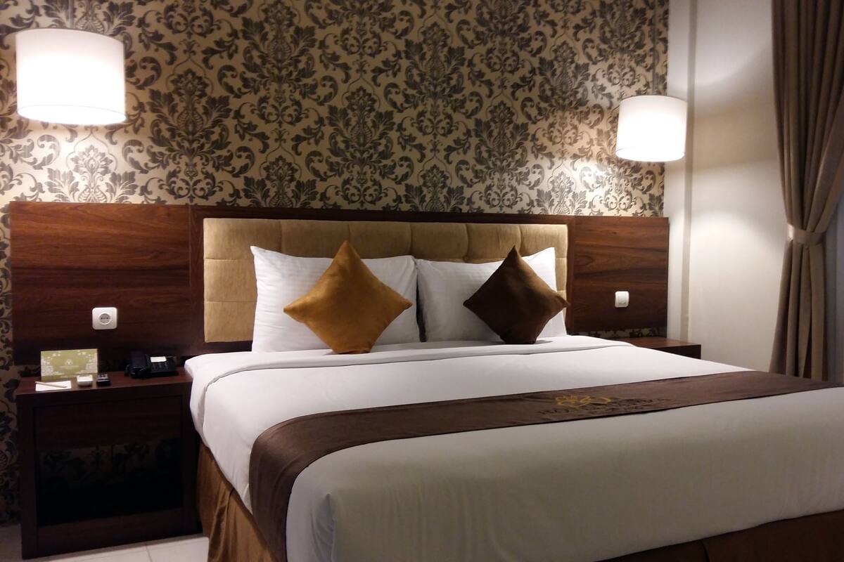 A hotel room with a large bed, two bedside tables, a wooden headboard, patterned wallpaper, and two wall-mounted lamps.