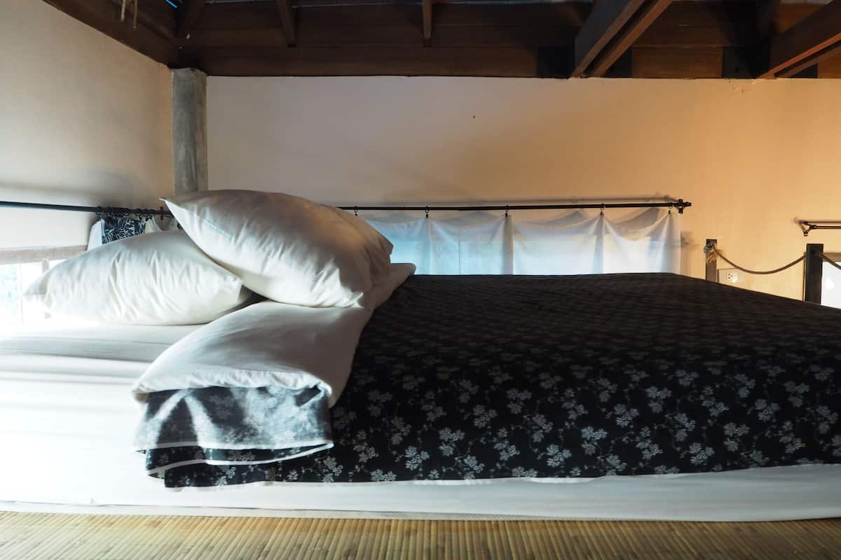 A bed with a black floral bedspread and white pillows, a wooden floor, and a simple, unadorned ceiling.