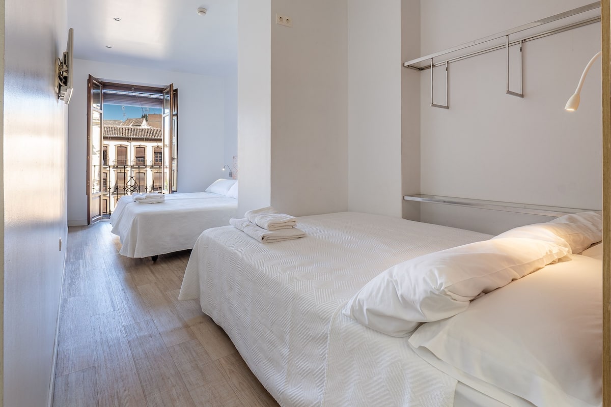 A hotel room with two beds, a wooden floor, and a view of buildings through an open door.