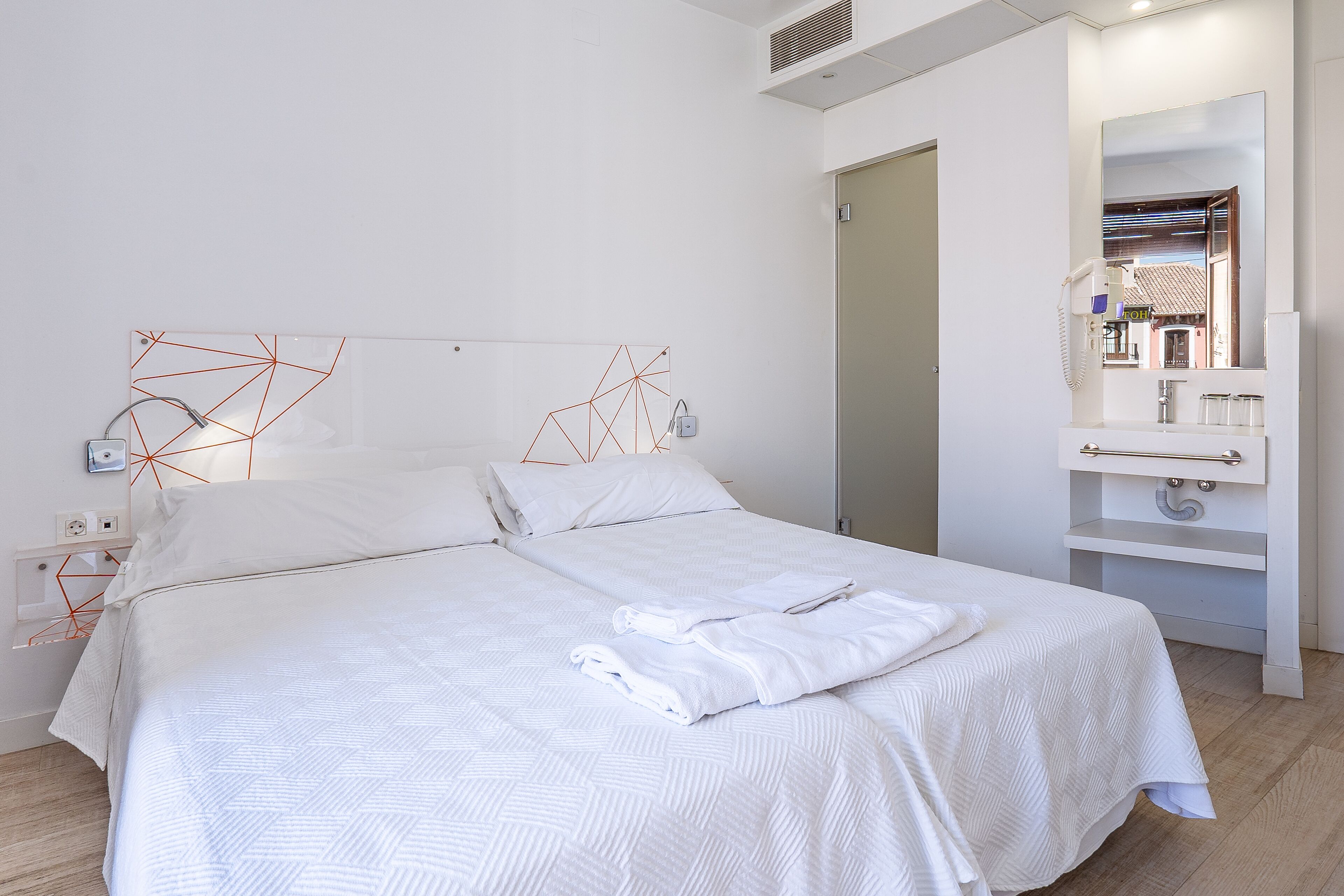 A neatly made bed with white linens and a folded towel, a bedside table with a lamp, and a bathroom with a mirror and sink.