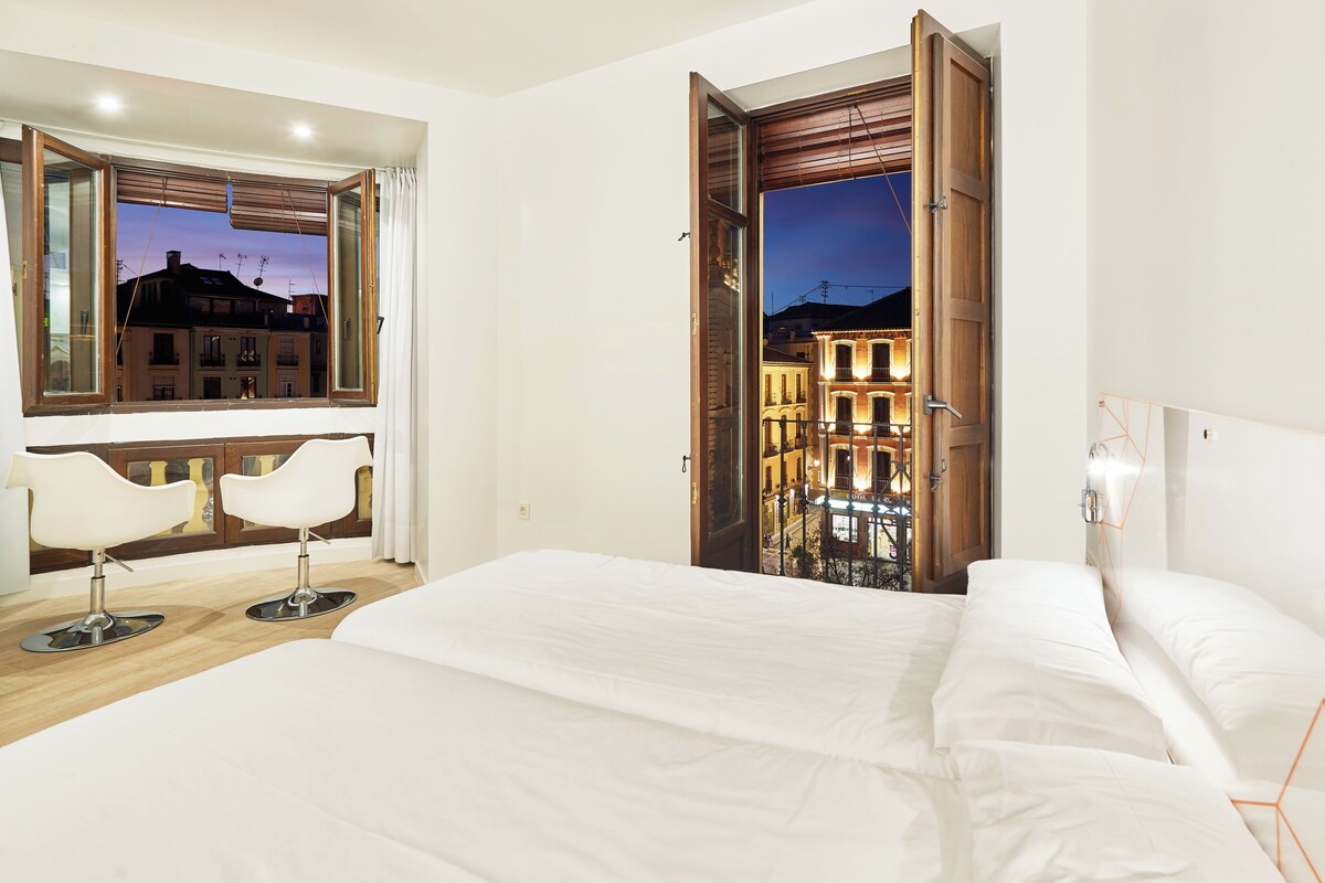 A hotel room with a large bed, two white chairs, a wooden door leading to a balcony, and a view of buildings at dusk.