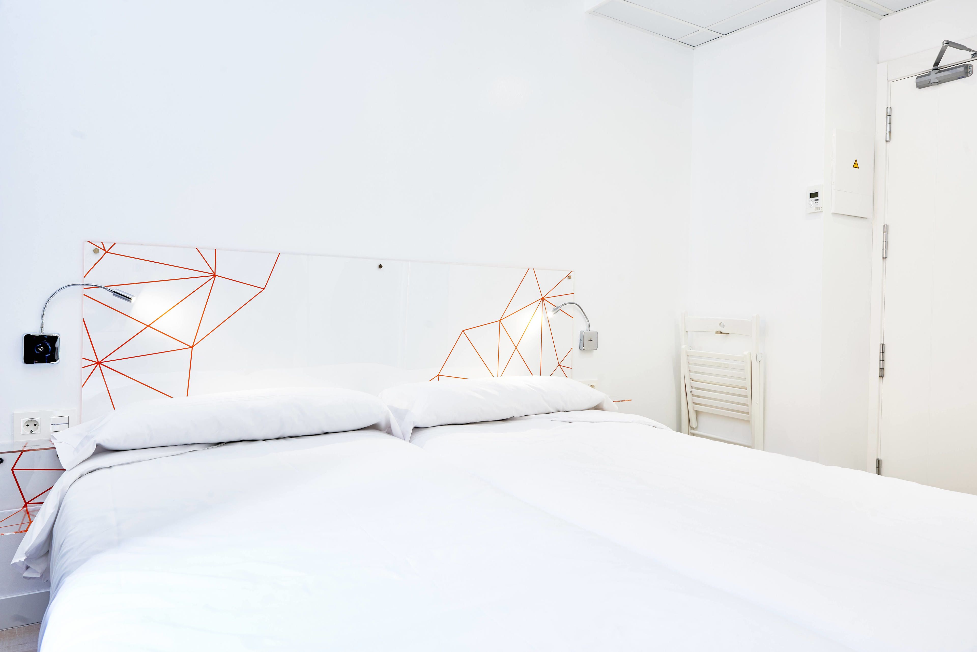 A modern hotel room with a neatly made bed, a bedside table with a lamp, and a wall-mounted control panel.