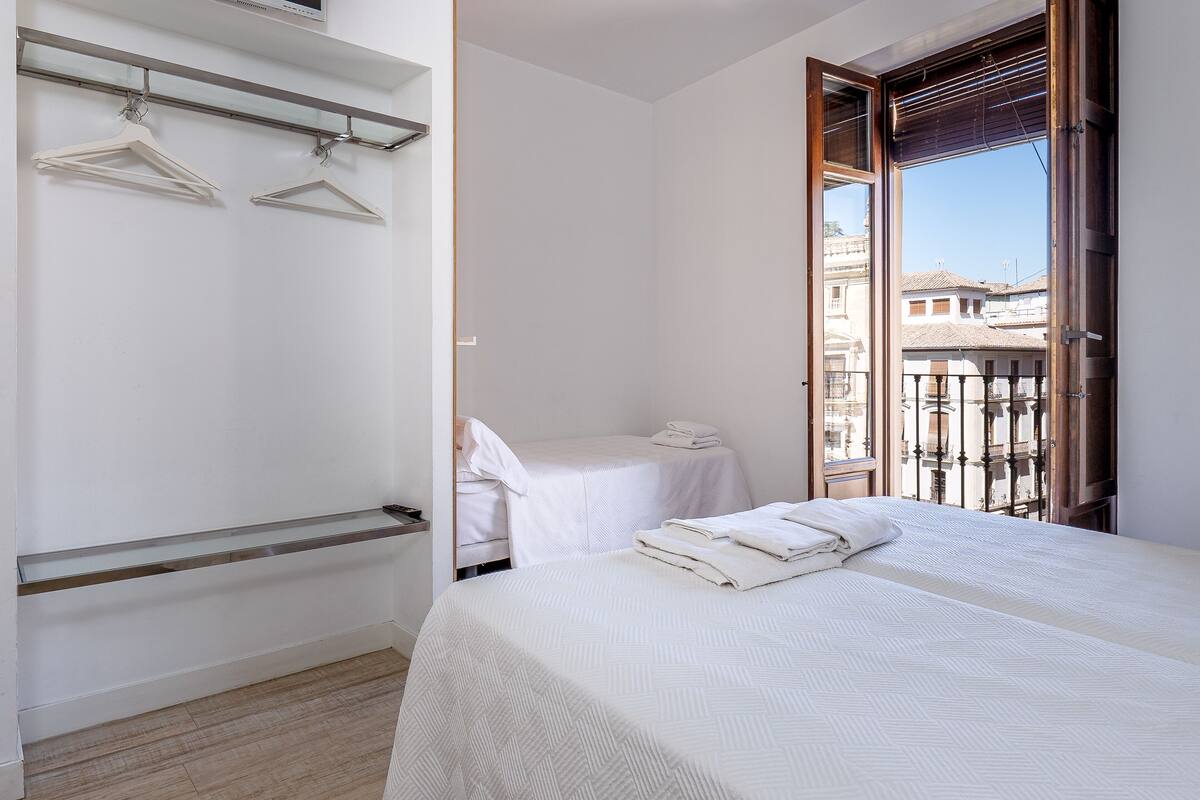 A bedroom with a bed, a bedside table, a hanging clothes rack, and a window with a view of buildings.