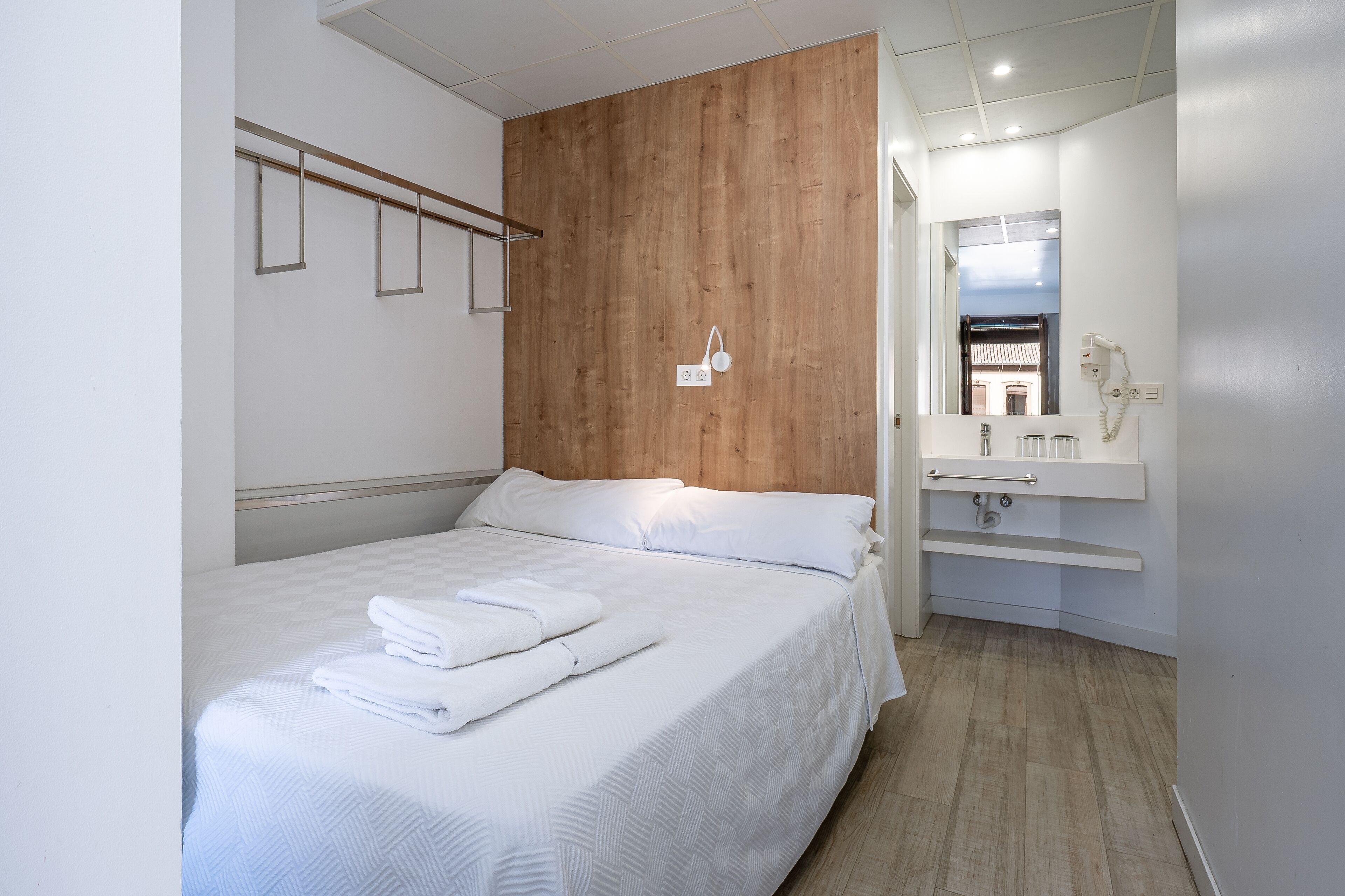 Habitación de hotel con cabecera de madera, cama con ropa de cama blanca, baño con lavamanos y espejo, y suelo de madera.
