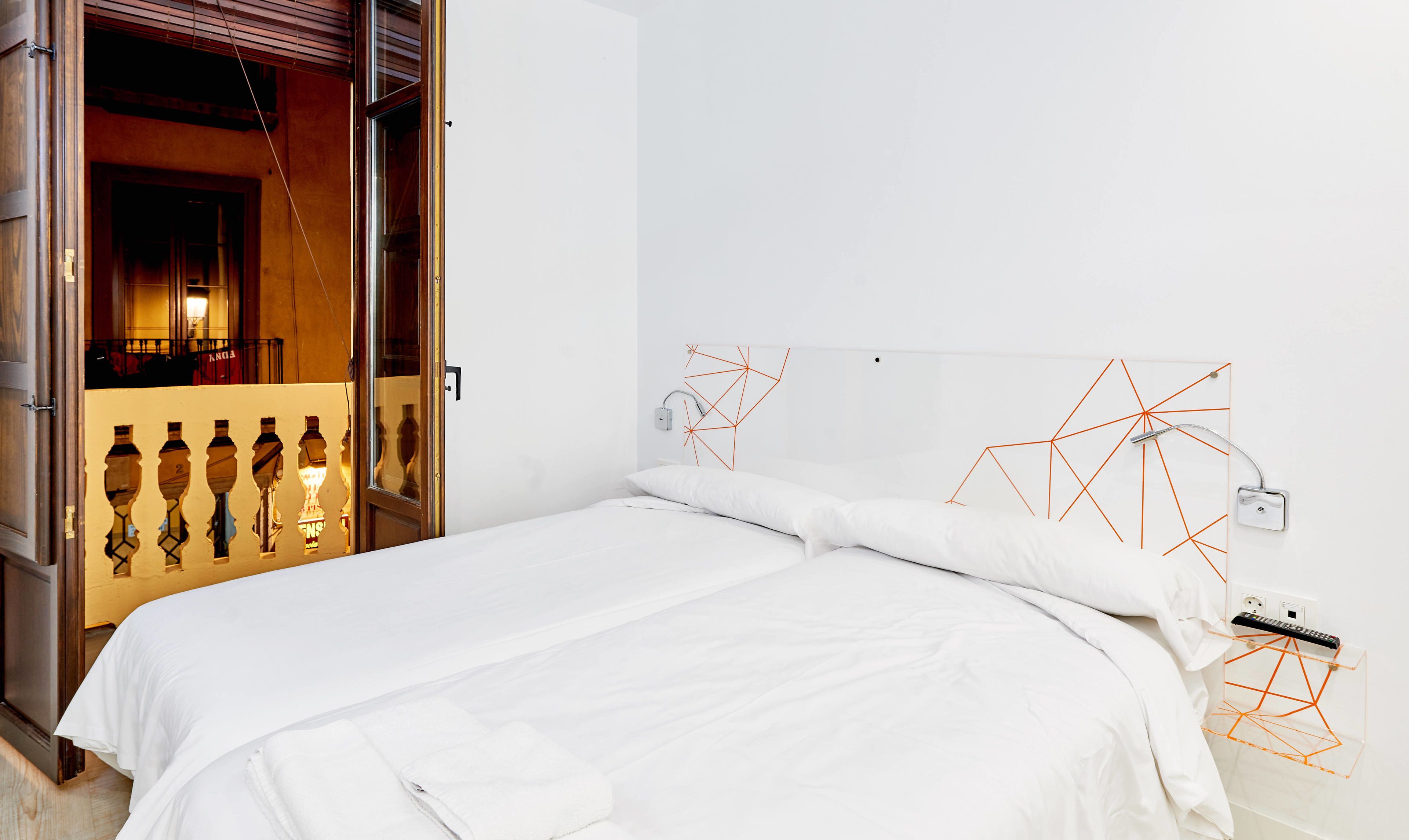 Habitación de hotel moderna con cama, ropa de cama blanca, una obra de arte geométrica en la pared y una puerta visible que conduce a otra habitación.